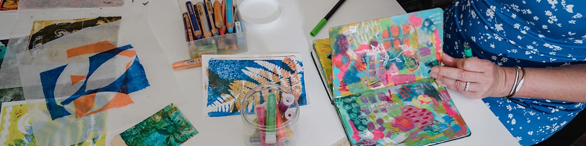 A person in a blue patterned dress creates colorful abstract art in a sketchbook on a white table, surrounded by art supplies, oil pastels, and other painted papers.