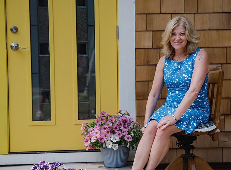 A woman with blonde hair in a blue floral dress sits on a chair by a yellow door, smiling. A pot of pink flowers is on the ground next to her, and the house has wooden siding.