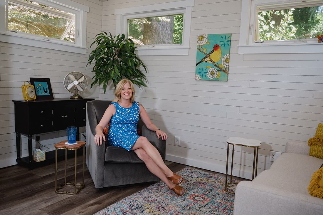 A woman in a blue floral dress sits and smiles in a gray armchair in a bright living room with white paneled walls, colorful decor, and large windows showing greenery outside.