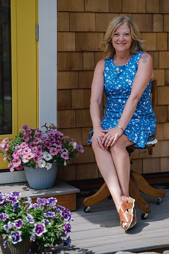 A woman in a blue floral dress sits on a wooden stool outside a house with wood siding and a yellow door. She smiles at the camera, and potted pink and purple flowers are nearby.