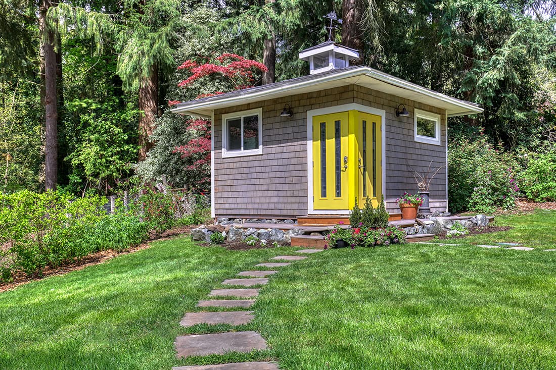A small gray cottage with a bright yellow double door sits in a lush green yard, surrounded by trees and bushes, with a stone path leading up to the entrance.