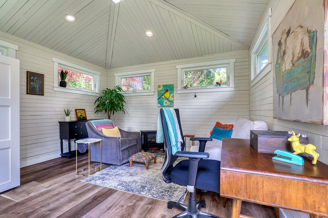 Bright, cozy home office with a vaulted ceiling, modern desk, office chair, gray armchair, indoor plants, vibrant artwork, and windows letting in natural light. The room features wood floors and stylish decor.